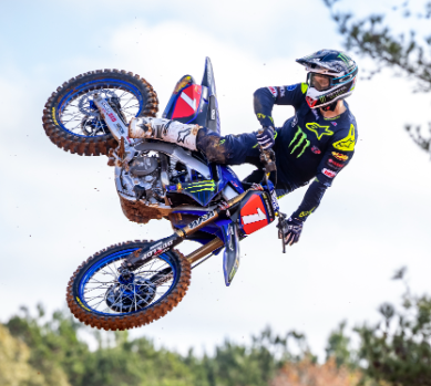 A motocross rider, wearing a Monster Energy racing suit and helmet, executes a daring midair maneuver on his Yamaha dirt bike, showcasing skill and athleticism against a backdrop of a partly cloudy sky and distant trees.