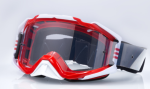 The image shows a pair of red and white motocross goggles. They have a large, clear lens and a strap. The goggles are angled slightly, and are displayed against a white background.