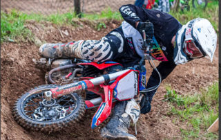 The image shows a motocross rider, wearing full protective gear, in a low position on a red and white dirt bike. The rider appears to be navigating a turn on a dirt track. The bike is partially buried in the loose dirt, suggesting a fall or near-miss. The overall impression is one of action and intensity within a motocross competition.