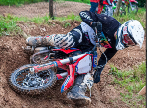 The image shows a motocross rider, wearing full protective gear, in a low position on a red and white dirt bike. The rider appears to be navigating a turn on a dirt track. The bike is partially buried in the loose dirt, suggesting a fall or near-miss. The overall impression is one of action and intensity within a motocross competition. 