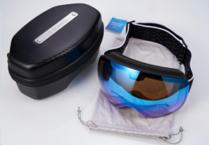 The image shows a pair of blue and black ski goggles with a black carbon fiber hard case and a grey drawstring bag. The goggles are resting inside the drawstring bag, and the case is positioned next to it. A small informational tag is also visible near the goggles. The overall presentation suggests a product shot, perhaps for an online store or catalog. 