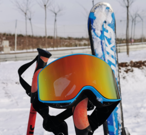 In the photo, a pair of ski goggles with orange lenses hangs between a red ski pole and a pair of blue skis. In the background is a snowy winter scene with bare trees and snowy ground visible. The picture is simple, highlighting the ski goggles, creating a winter sports atmosphere.