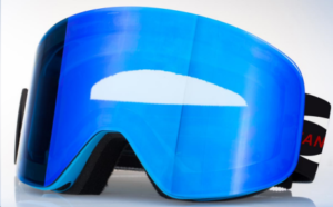 The image shows a pair of blue ski goggles. The lenses are a vibrant blue with a slight mirror effect, and the frame is a teal or light blue color. A portion of a black strap with red lettering is visible. The goggles are displayed against a plain white background. 